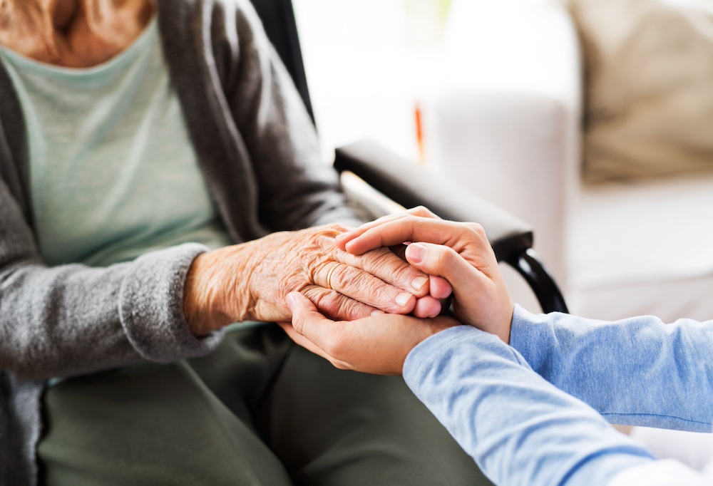 A senior woman sitting in a wheelchair holds hands with a caregiver